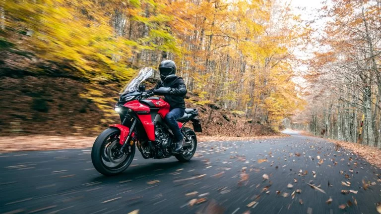 rood met zwarte Yamaha Tracer 9 rijd in de herfst door een bosrijke omgeving te zien vanuit het voorzijaanzicht met bladeren op de weg