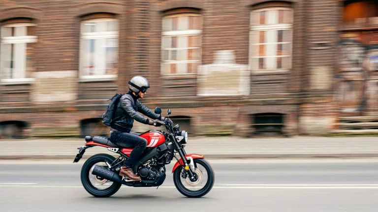 Actiefoto 2023 Yamaha XSR 125 Red Line op afstand met motorrijder met rugzak op die langs een oud pakhuis rijdt