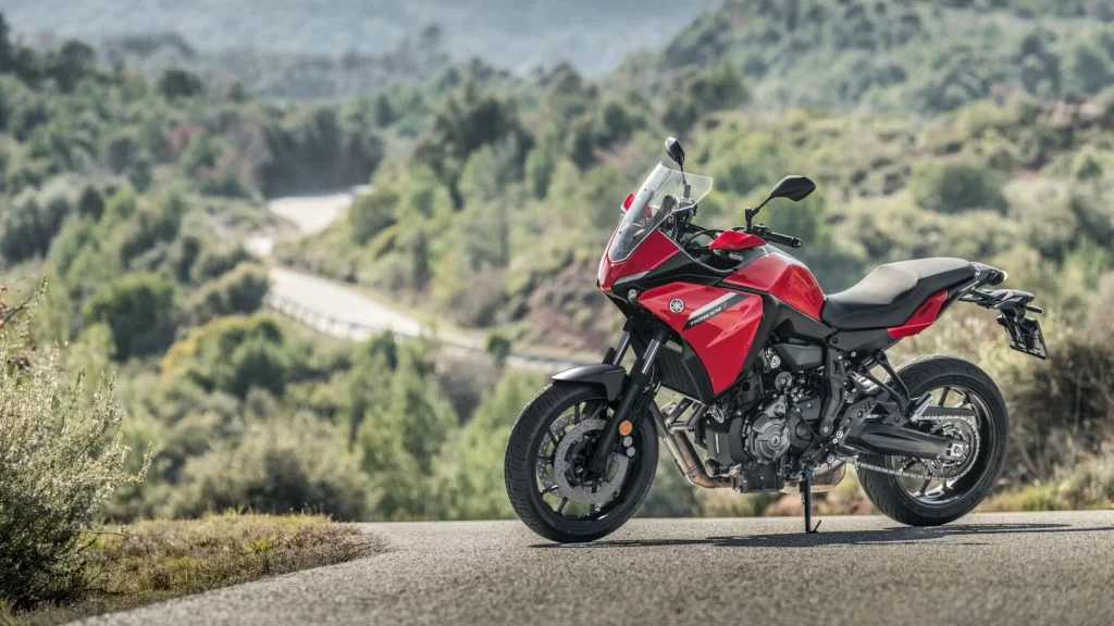 rode Yamaha Tracer 7 te zien vanuit het zijaanzicht staat op een weg met als achtergrond een dal en een bergachtige omgeving