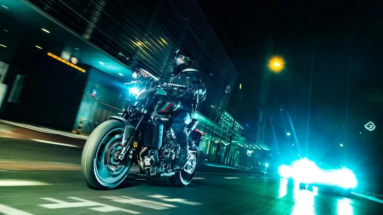 zwarte Yamaha MT-09 met turquoise velgen rijd in het donker met gebouwen en auto's op de achtergrond te zien vanuit het zijaanzicht