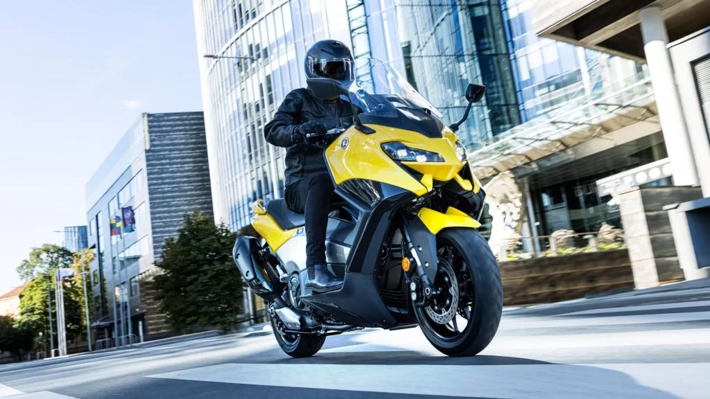 Actiefoto 2022-Yamaha-XP500A-EU-Extreme_Yellow-Action-003 met motorrijder die door de stad rijdt met groot gebouw rechts