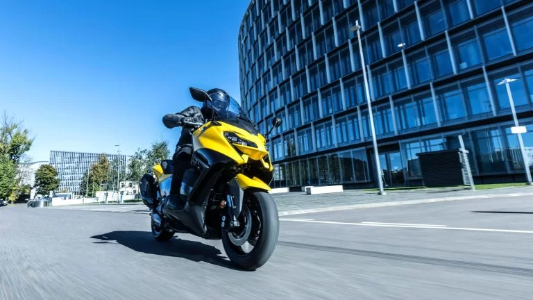 Actiefoto 2022-Yamaha-XP500A-EU-Extreme_Yellow-Action-003 met motorrijder die door de stad rijdt met groot gebouw rechts