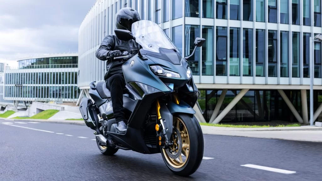 2022-Yamaha-XP500ADX-EU-Dark_Petrol-Action vooraanzicht met motorrijder die hangend door de bocht rijdt met modern kantoorgebouw op de achtergrond