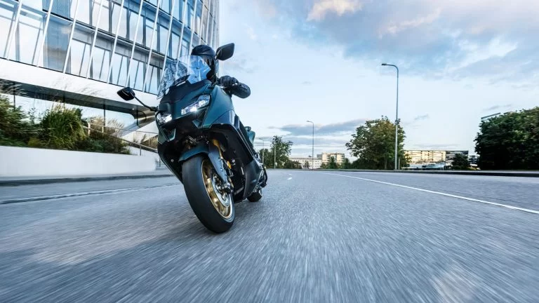 2022-Yamaha-XP500ADX-EU-Dark_Petrol-Action vooraanzicht met motorrijder die hangend door de bocht rijdt met rond kantoorgebouw links in beeld