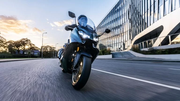 2022-Yamaha-XP500ADX-EU-Dark_Petrol-Action vooraanzicht met motorrijder die hangend door de bocht rijdt met modern kantoorgebouw rechtsin beeld
