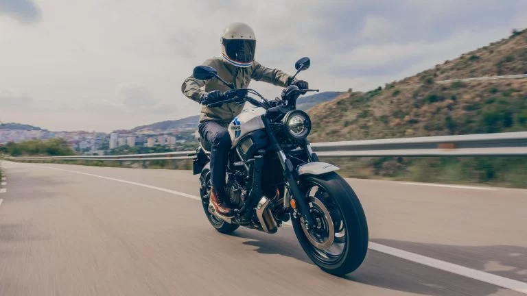 Actiefoto 2022-Yamaha-XS700-EU-Historic_White-Action-001-03 vooraanzicht van motorrijder die door heuvelachtig gebied rijdt met stad op de achtergrond