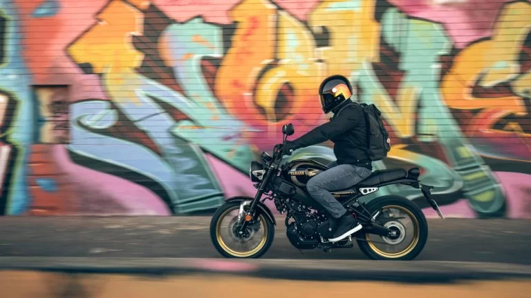 Actiefoto 2023-Yamaha-XS125SV-EU-Historic_Black-Action-004-03 met motorrijder met rugzak de langs een muur met graffiti rijdt.
