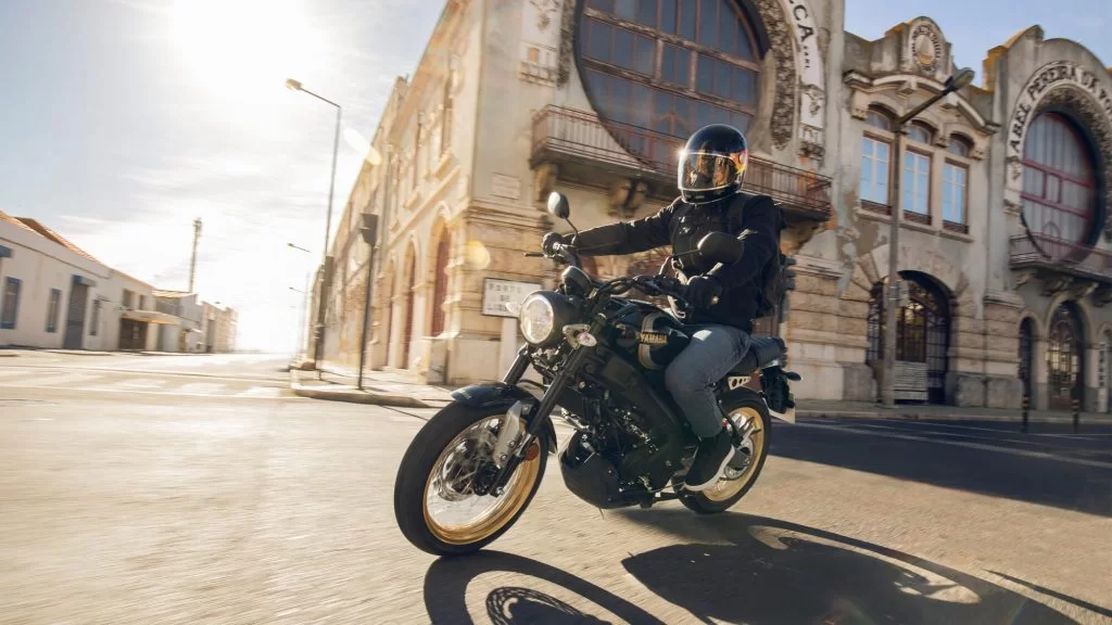 Actiefoto 2023-Yamaha-XS125SV-EU-Historic_Black-Action-004-03 met motorrijder die de bocht om gaat op rustige straat in een voorwijk van stad