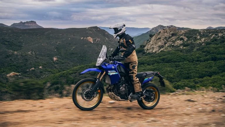 Motorrijder rijdt op een adventure motor over een onverharde weg met bergen en groene bosjes op de achtergrond