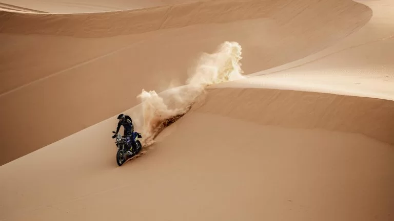 afstandsshot met motorrijder die over de duinen heenrijdt en stof doet opwaaien op een adventure motor