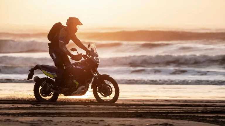 Adventure motor met motorijder die over het strand rijdt met de zee en ondergaande zon op de achtergrond