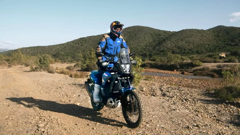 motorrijder rijdt op een blauwe adventure motor over een onverhard pad met bergen op de achtergrond