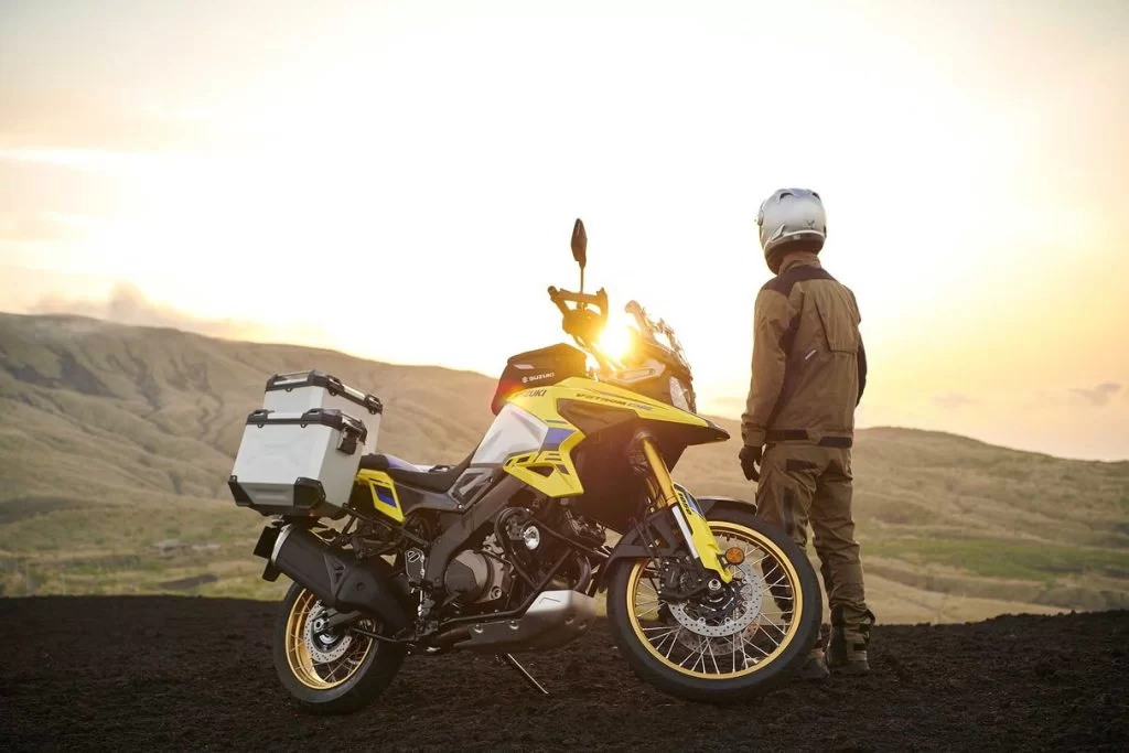 Lifestylefoto 2023 Suzuki V Strom 1050 DE met motorrijder die naast de motorfiets staat uitkijkend over vallei met zonsondergang op achtergrond