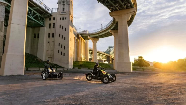 twee Can-Am Rykers 900 rijden onder een rond bruggebouw door met grote pilaren te zien vanuit het voorzijaanzicht met de zonsondergang op de achtergrond